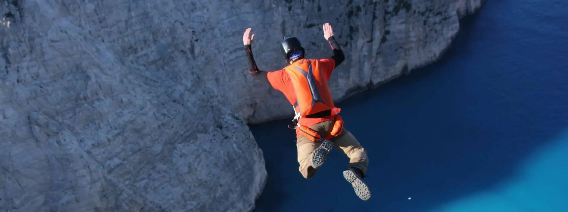 A base jumper leaps off a cliff seen from above, he wears an orange parachute manufactured by adrenalin base.